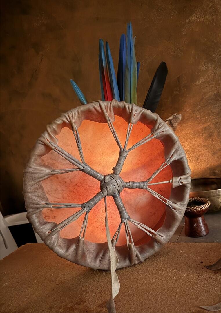 Eine handgefertigte Trommel mit durchscheinendem, gespanntem Fell, das auf der Rückseite Schnürungen und Knoten aufweist. Bunte Federn - blau, rot und schwarz - stehen aufrecht hinter der Trommel vor einem braunen Hintergrund.