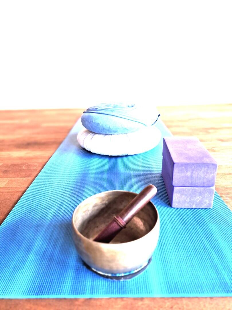Eine Yogamatte, davor eine Klangschale und ein Schlägel, zwei violette Yogablöcke und zwei übereinander gestapelte Meditationskissen auf einem Holzboden.