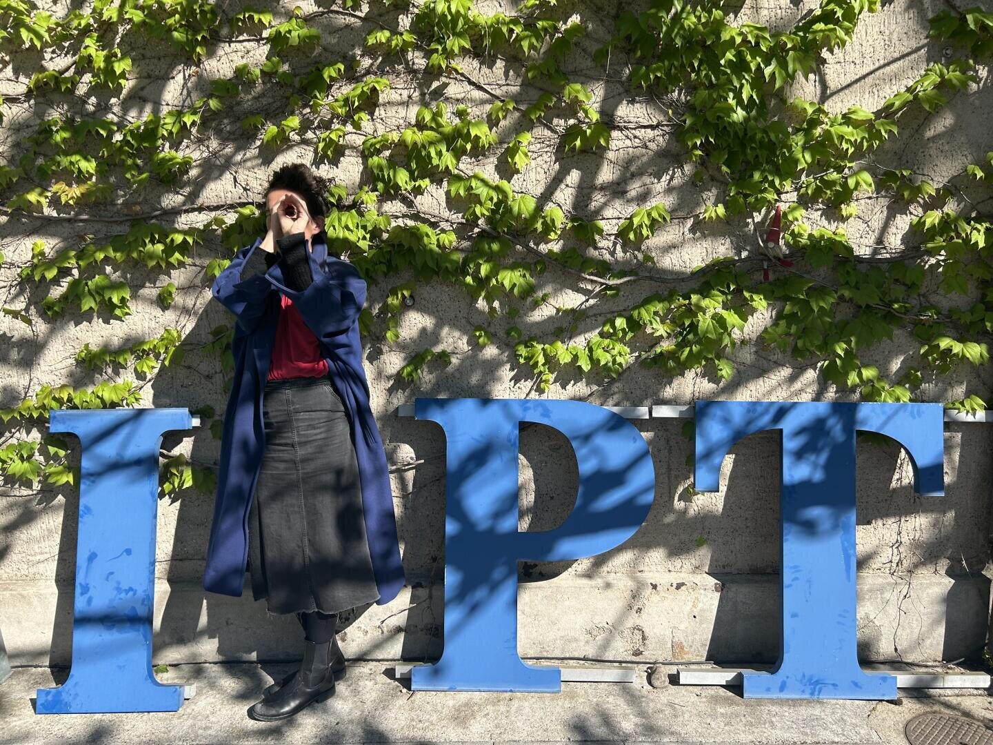 Eine Person steht neben großen blauen Buchstaben, die „IPT“ bilden, vor einer sonnenbeschienenen, mit Efeu bewachsenen Wand und formt mit den Händen einen Kreis um ein Auge.