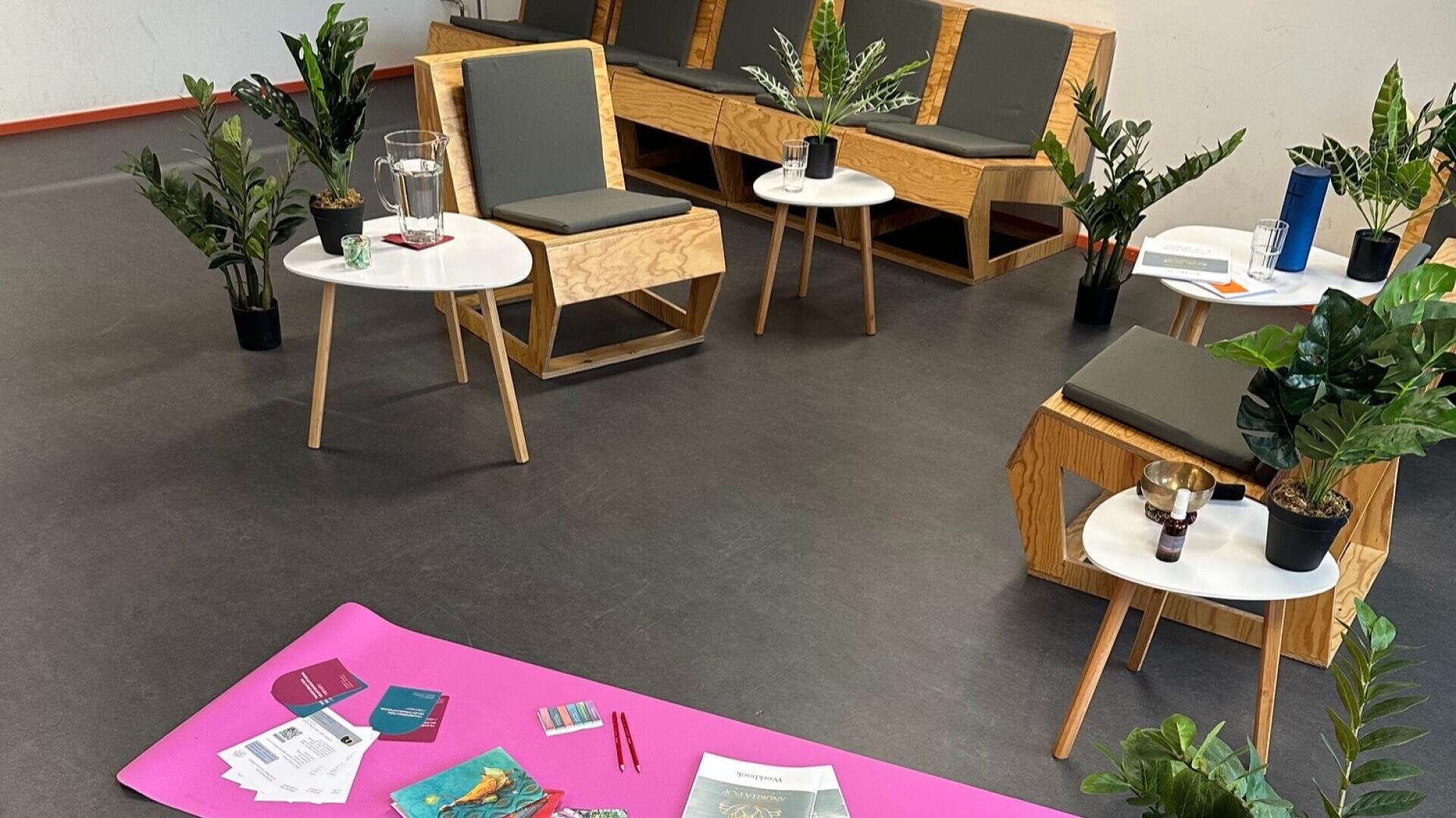Ein Raum mit Holzstühlen, kleinen Tischen, auf denen Pflanzen und Gläser stehen, und einer rosa Matte auf dem Boden, auf der Zeitschriften, Broschüren, Karten und Stifte liegen. Die Einrichtung ist ungezwungen, und hier und da stehen ein paar Topfpflanzen.