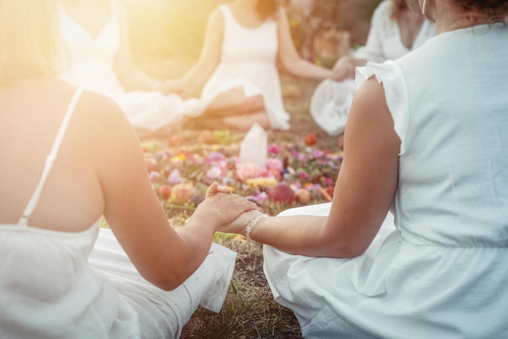 Weiß gekleidete Frauen sitzen im Freien in einem Kreis und halten sich an den Händen um ein buntes Arrangement aus Blumen und Kristallen auf dem Boden, während das Sonnenlicht die Szene beleuchtet.
