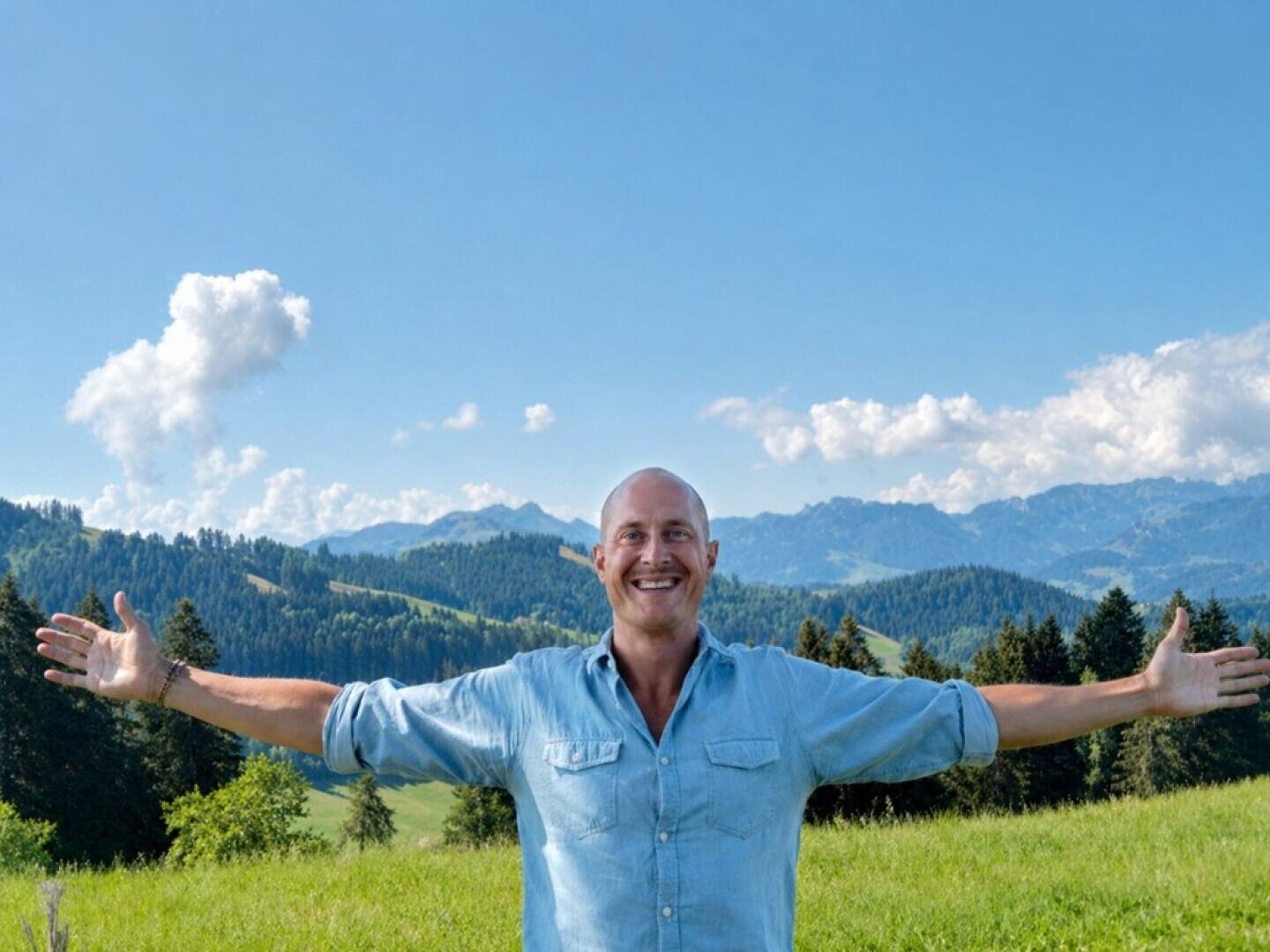 Ein lächelnder Mann mit ausgestreckten Armen, der auf einer Wiese steht, im Hintergrund sind Berge zu sehen.
