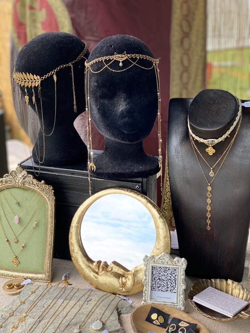 Eine Schmuckauslage zeigt goldene Kopfschmuckstücke auf schwarzen Ständern, Halsketten, Ringe, einen Spiegel mit goldenem Zierrahmen, eine Halskettenablage aus grünem Samt sowie einen QR-Code auf einer Karte – alles auf einem strukturierten Tuch angeordnet.