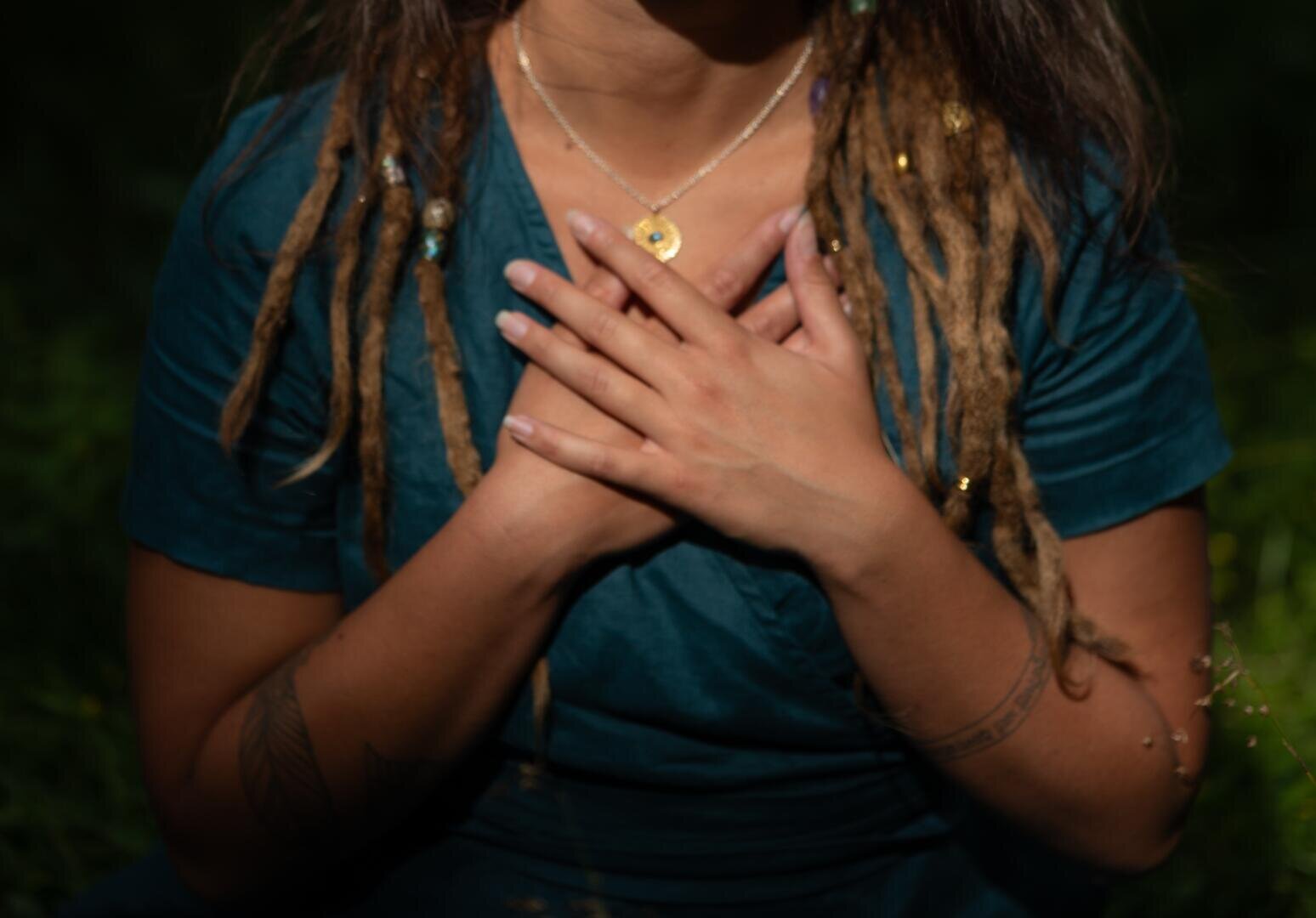 Eine Person mit langen, braunen Dreadlocks und Tätowierungen am Arm trägt ein blaugrünes Kleid und eine Halskette und sitzt im Freien, die Hände vor der Brust verschränkt.