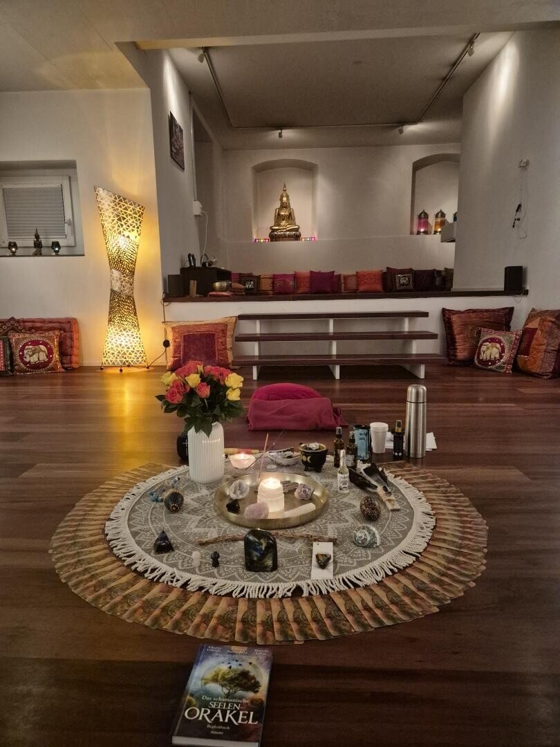 Ein gemütlicher, sanft beleuchteter Meditationsraum mit einer Buddha-Statue auf einem erhöhten Podest, bunten Kissen an den Wänden und einer kreisförmigen Anordnung von Kerzen, Kristallen, Ölen und Tarotkarten auf dem Boden, daneben Blumen und ein Buch.