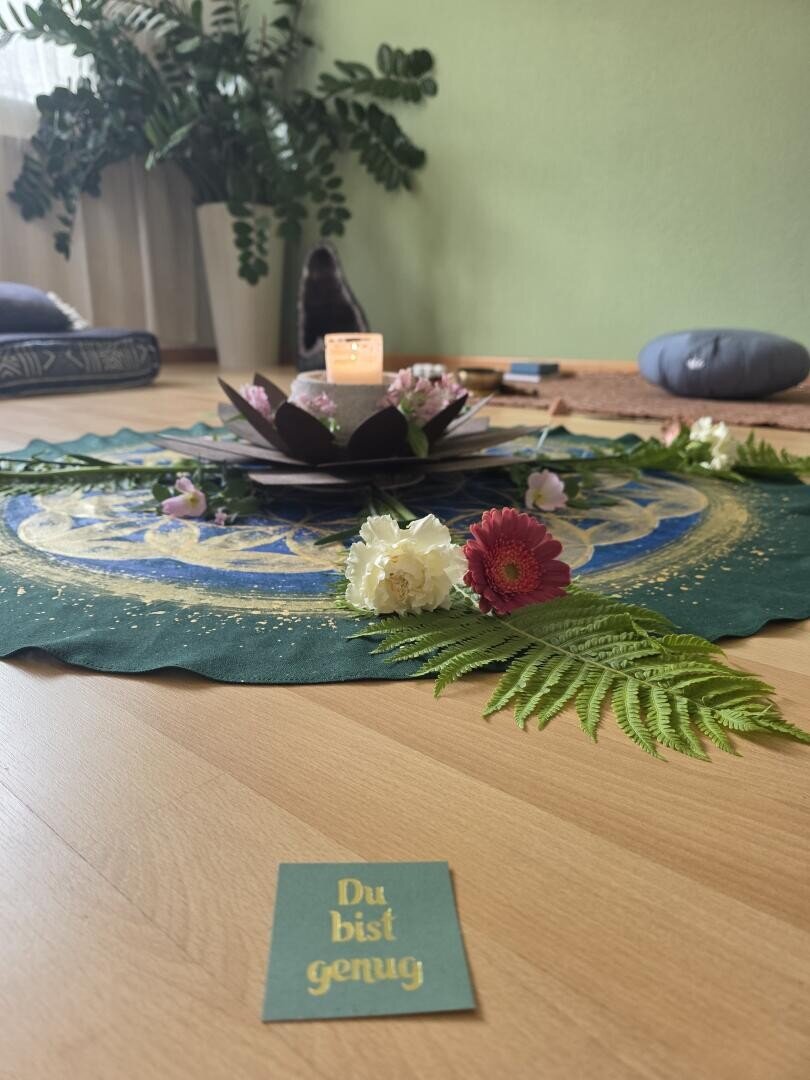 Auf dem Boden steht ein harmonisches Arrangement auf einem grünen Tuch mit Kerzen, Blumen und Farnen; auf einer Karte im Vordergrund steht auf Deutsch „Du bist genug“.