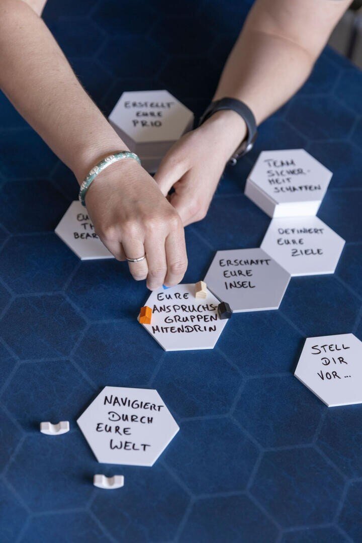 Die Hände einer Person ordnen weiße sechseckige Fliesen mit handgeschriebenen deutschen Sätzen auf einer blau gemusterten Oberfläche an. Die sichtbaren Kacheln enthalten Wörter wie "Eure Anspruchsgruppen Mittendrin" und "Navigiert durch eure Welt".
