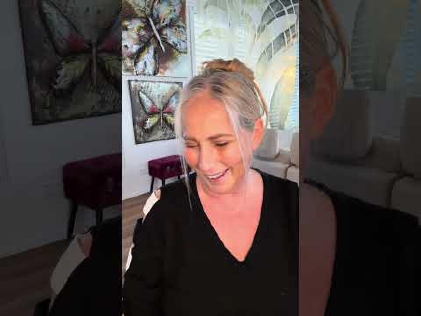 Eine Frau mit zurückgebundenen grauen Haaren und schwarzem Oberteil sitzt lächelnd in einem gut beleuchteten Raum, in dem im Hintergrund Schmetterlingskunstwerke und modernes Dekor zu sehen sind.