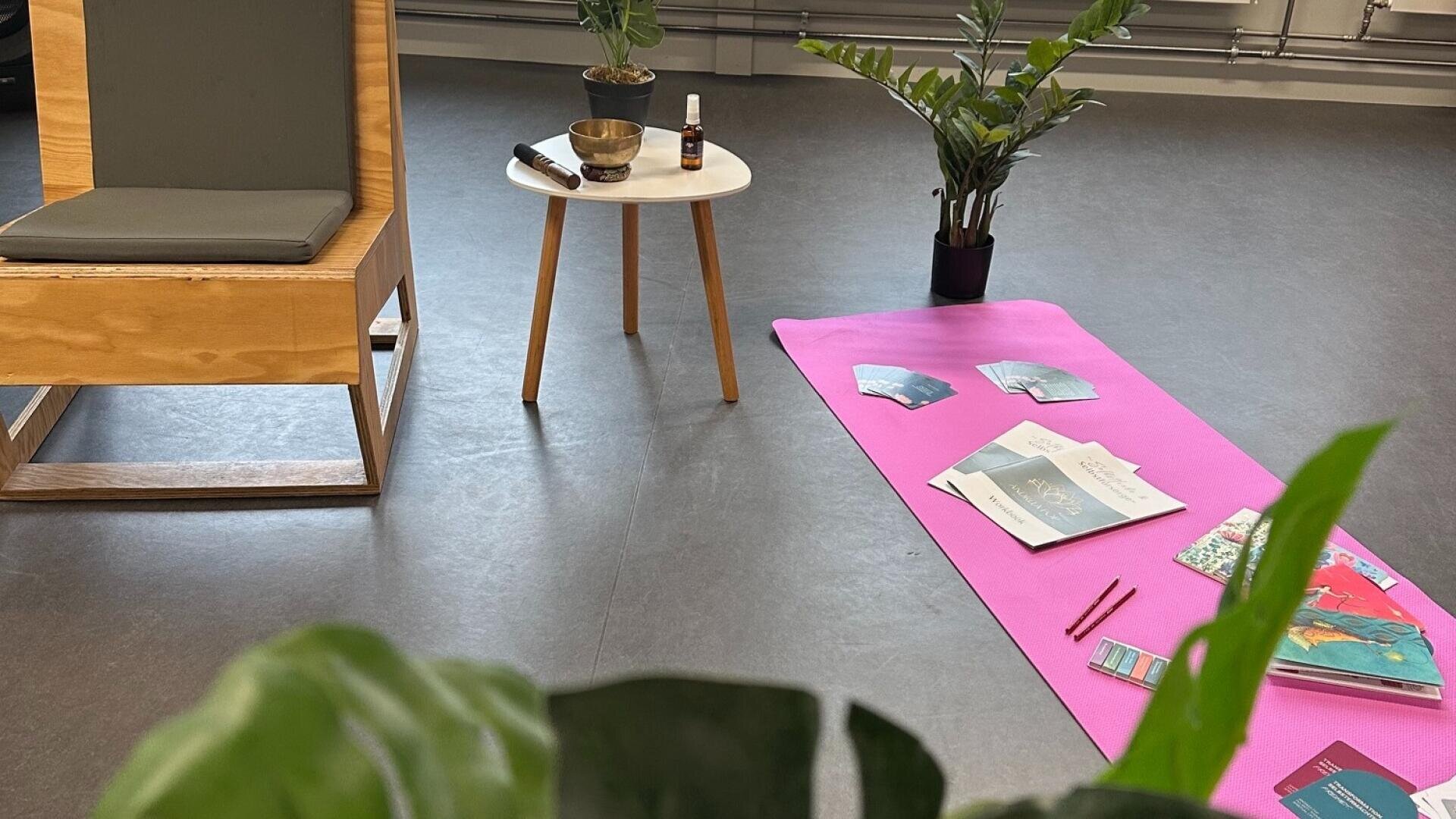 Auf dem Boden liegt eine rosa Yogamatte mit Büchern, Karten und Stiften, daneben steht ein kleiner Tisch mit einer Schale und einer Sprühflasche. In der Nähe stehen ein Holzstuhl und Topfpflanzen. Die Umgebung wirkt ruhig und einladend.