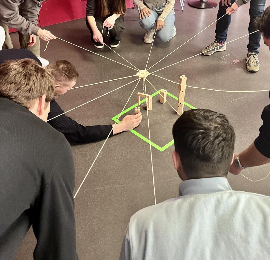 Eine Gruppe von Menschen kniet in einem Kreis, jeder hält eine Schnur, die mit einer Holzstruktur in der Mitte verbunden ist, und arbeitet gemeinsam an einer teambildenden Maßnahme auf einem dunklen, mit grünem Klebeband markierten Boden.