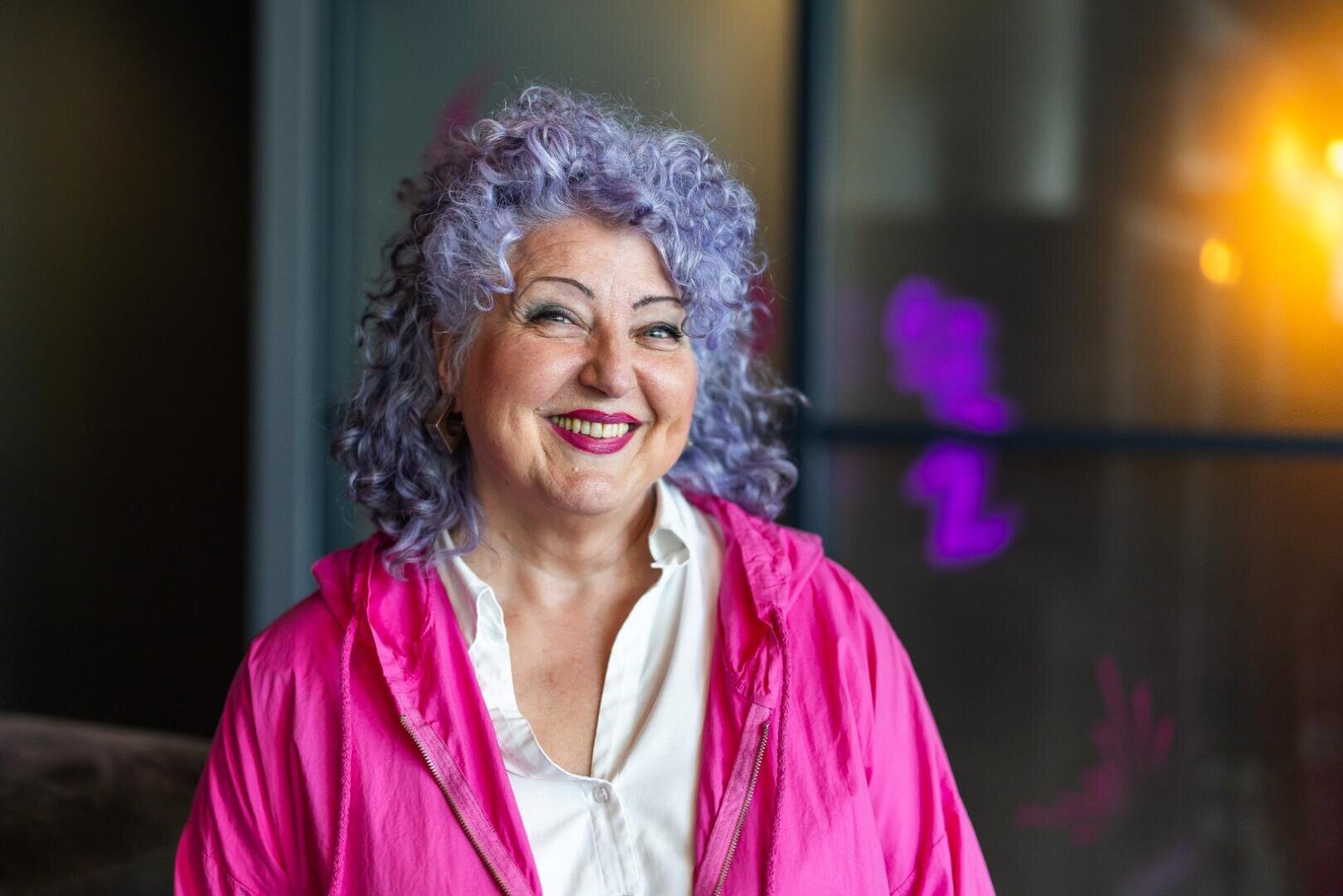 Lächelnde ältere Frau mit lockigem lavendelfarbenem Haar, die eine leuchtend pinke Jacke und ein weißes Hemd trägt. Sie steht in einem Innenraum mit einem unscharfen, modernen Hintergrund und weicher Beleuchtung.