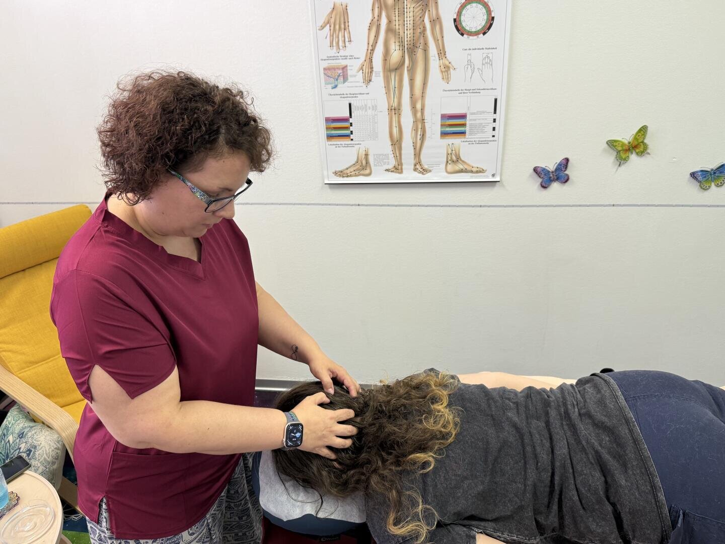 Eine Massagetherapeutin in einem weinroten OP-Oberteil führt bei einer Klientin, die mit dem Gesicht nach unten auf einer Behandlungsliege liegt, eine Kopfmassage durch.