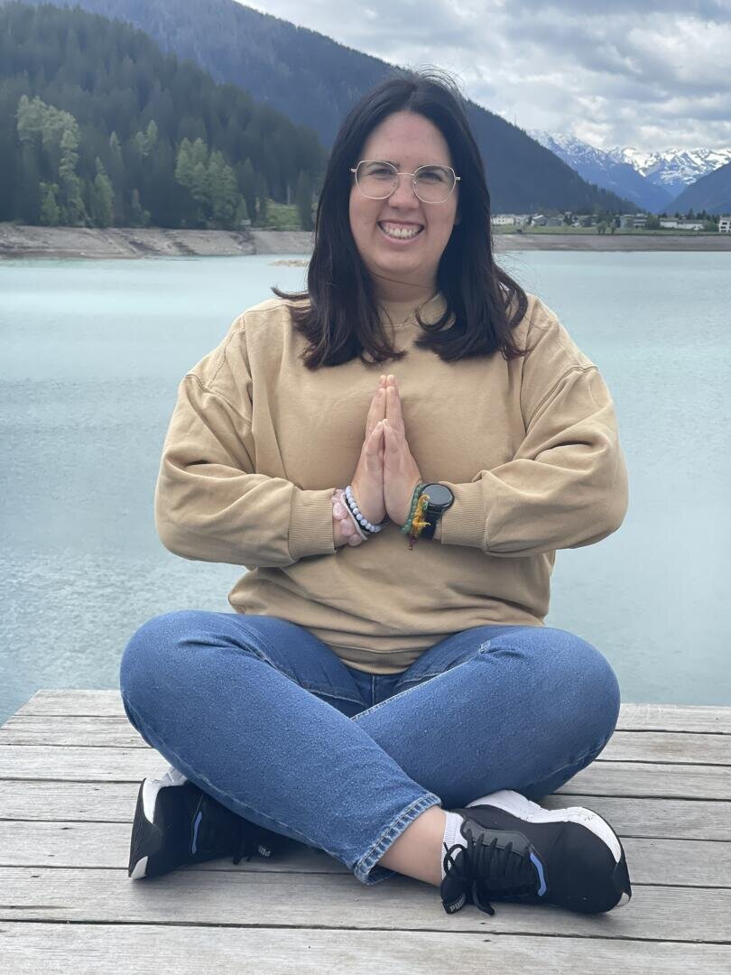 Eine Frau mit langen dunklen Haaren, Brille und beigem Sweatshirt sitzt im Schneidersitz auf einem hölzernen Steg an einem türkisfarbenen See, lächelt und hält die Hände in Gebetshaltung. Im Hintergrund sind Berge, Bäume und Wolken zu sehen.