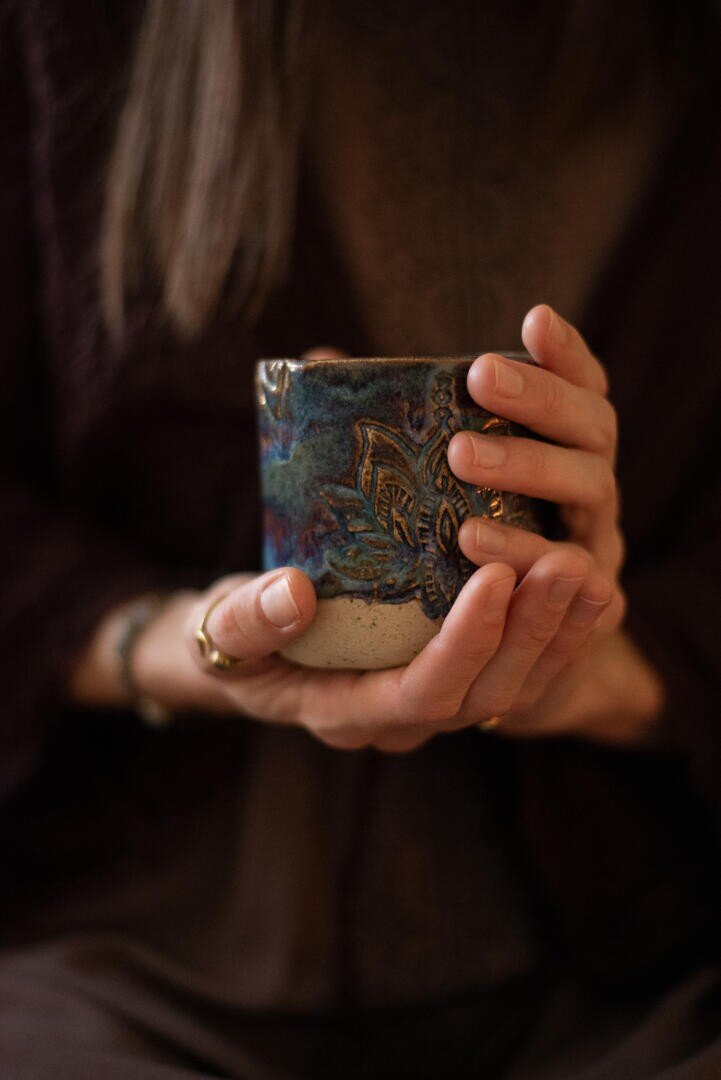 Eine Person mit langen Haaren hält mit beiden Händen eine handgefertigte Keramik-Tasse fest, wobei ihre Finger sich sanft um den Becher legen. Die Tasse ist mit blauen und braunen Mustern verziert. Die Szene strahlt Wärme und Gemütlichkeit aus.