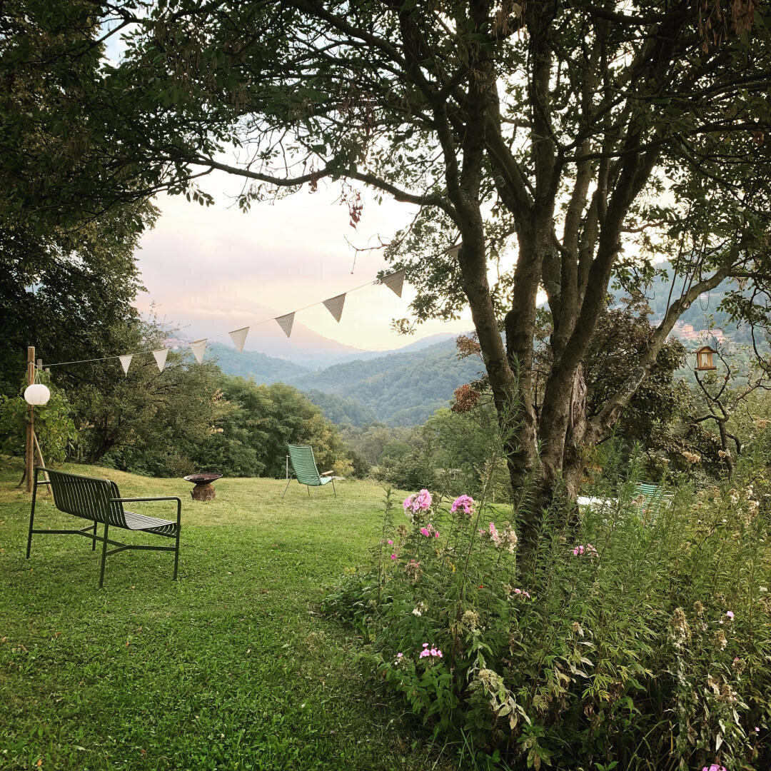 Eine friedliche Gartenszene mit einer Bank, Stühlen, Wildblumen und einem Baum, der mit dreieckigen Wimpelflaggen behängt ist, mit Blick auf grüne Hügel und Berge unter einem bewölkten Himmel.