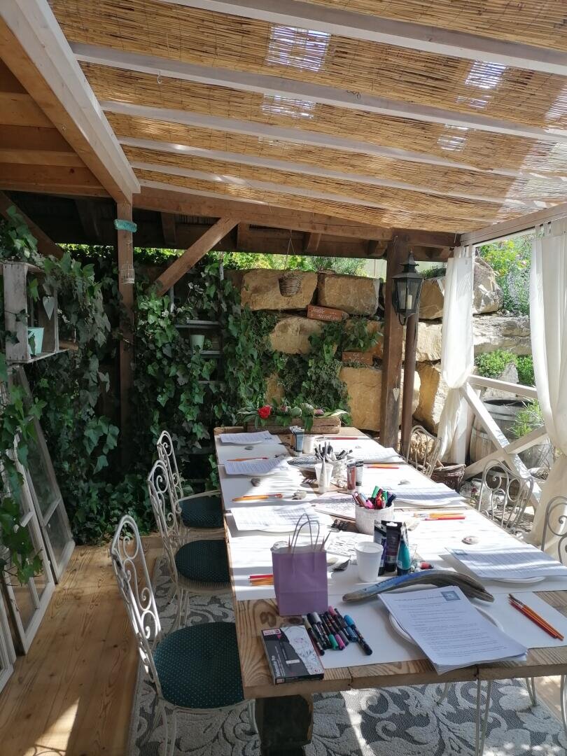 Ein sonnendurchfluteter Arbeitsbereich im Freien mit einem langen Tisch, auf dem Papiere, Stifte und Malutensilien liegen. Um den Tisch herum stehen mehrere Stühle; grüne Pflanzen und Kletterpflanzen schmücken den gemütlichen, schattigen Terrassenbereich.