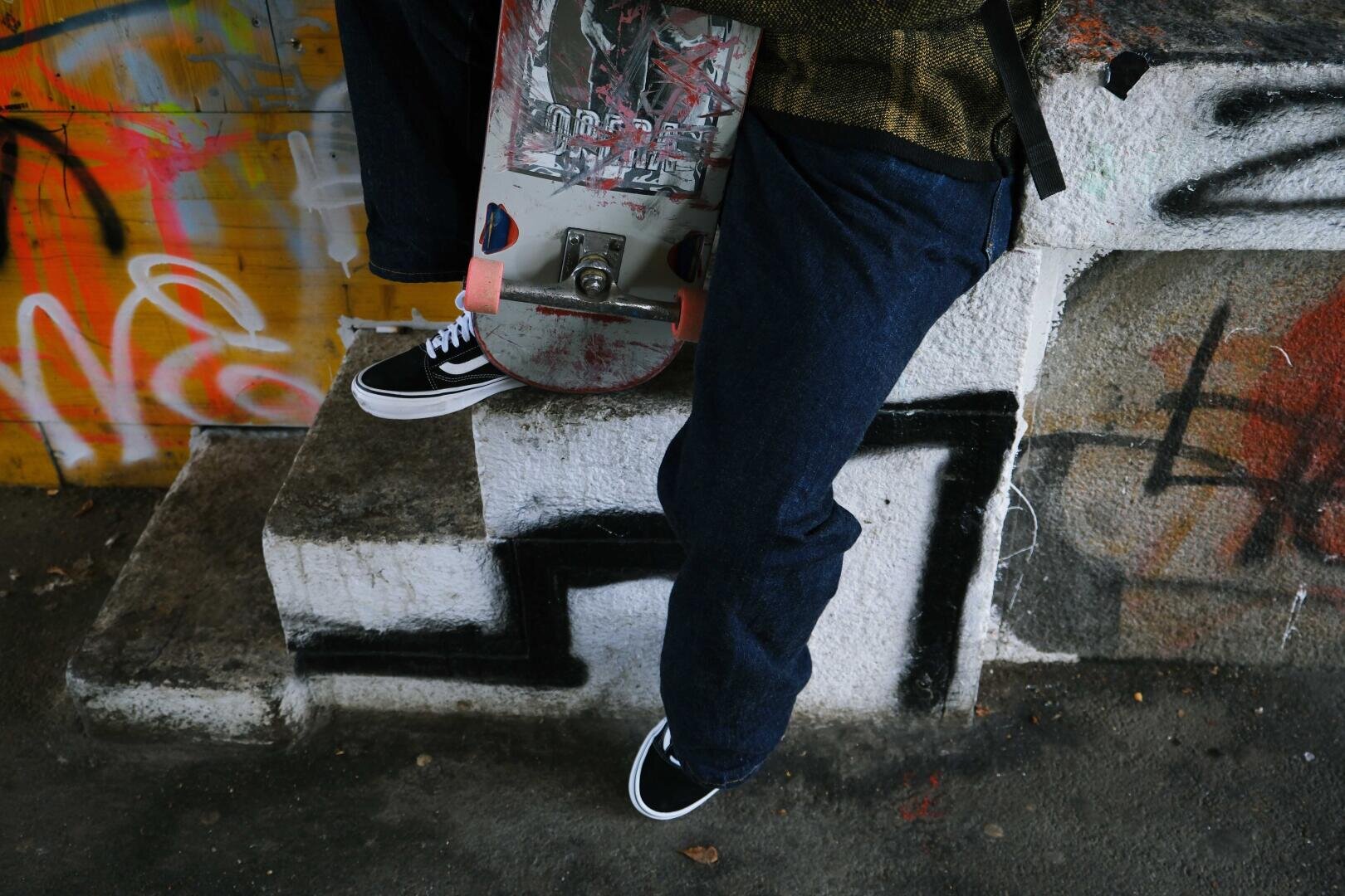 Eine Person in blauen Jeans und schwarzen Turnschuhen lehnt an einer Betonstufe und hält ein Skateboard in der Hand. Bunte Graffiti bedecken die Wand und die Stufen um sie herum.