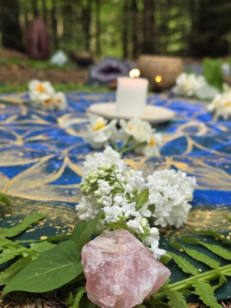 Eine Nahaufnahme eines rosa Kristalls, weißer Blumen und grüner Blätter, die auf einem blau-golden gemusterten Tuch arrangiert sind, mit einer brennenden Kerze und weiteren Blumen, die im Hintergrund verschwommen erscheinen – aufgenommen im Freien in einer Waldkulisse.