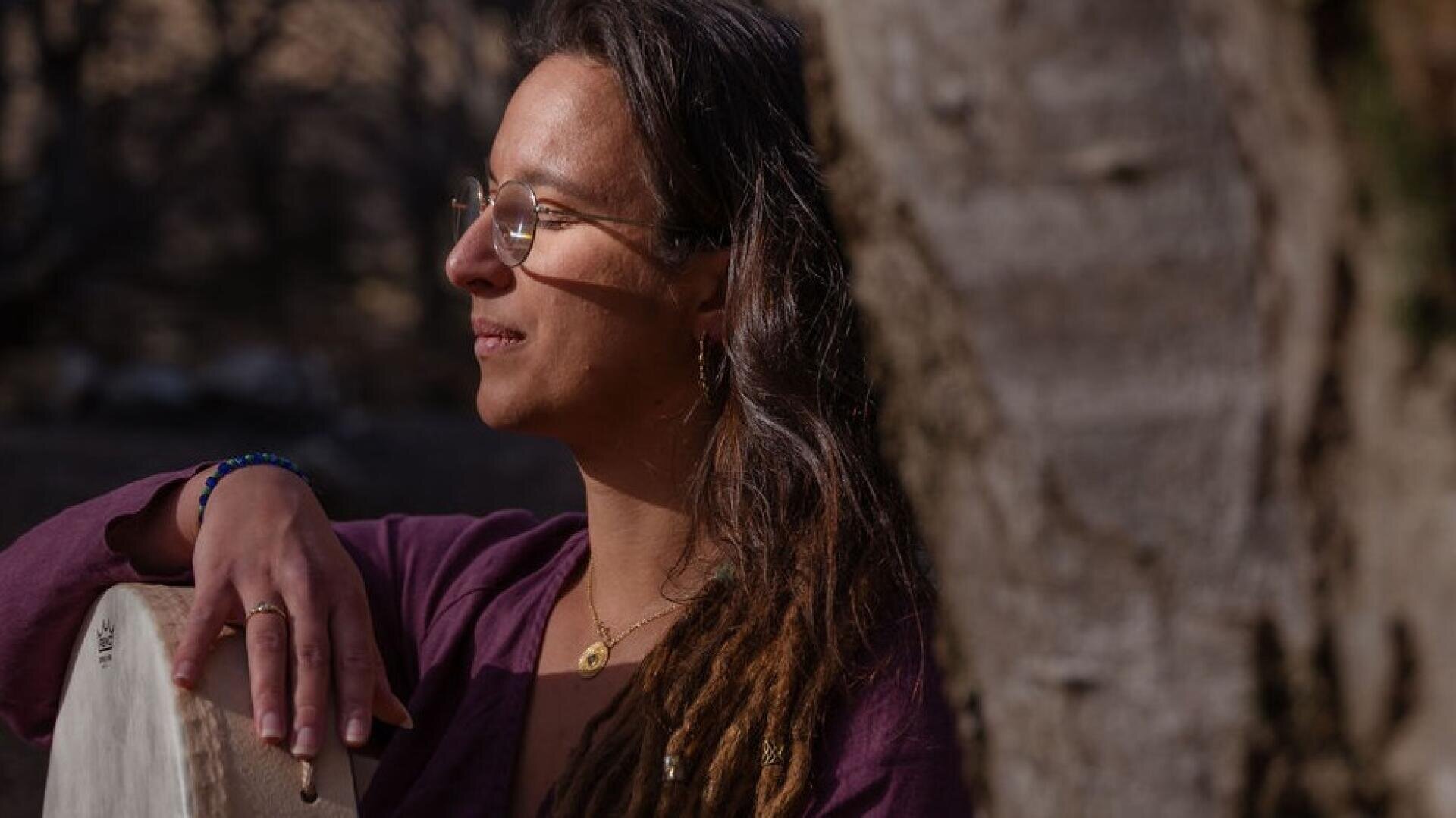 Eine Person mit langen, braunen Dreadlocks und Brille steht im Freien neben einem Baum, trägt eine lila Jacke und goldenen Schmuck, stützt sich mit der Hand auf eine Trommel und schaut nachdenklich in die Ferne.