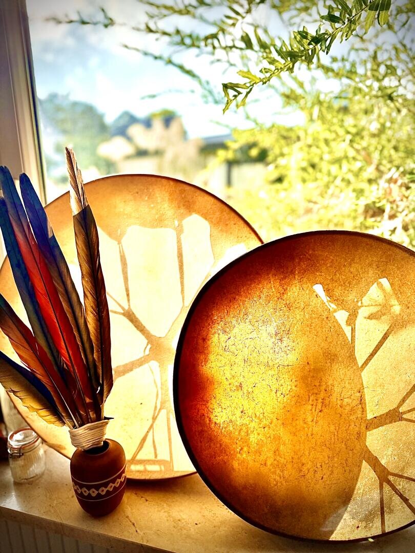 Zwei Handtrommeln mit durchscheinenden, sonnenbeschienenen Fellen stehen auf einer Fensterbank neben einer kleinen verzierten Vase mit großen, bunten Federn. Durch das Fenster sind helles Sonnenlicht und Grünzeug zu sehen.