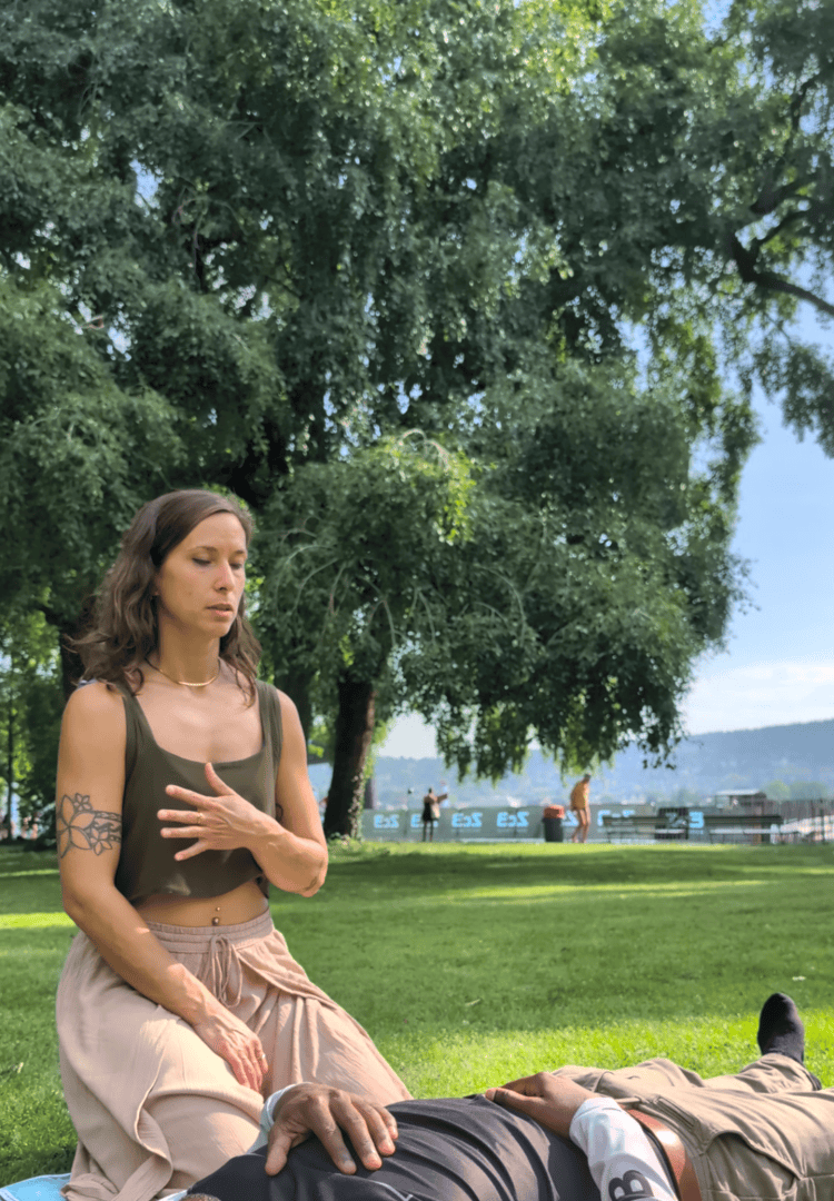 Eine Frau sitzt auf dem Rasen, die Hand auf der Brust, die Augen geschlossen, neben einer liegenden Person, in einem Park mit Bäumen und einem in der Ferne liegenden Gewässer.