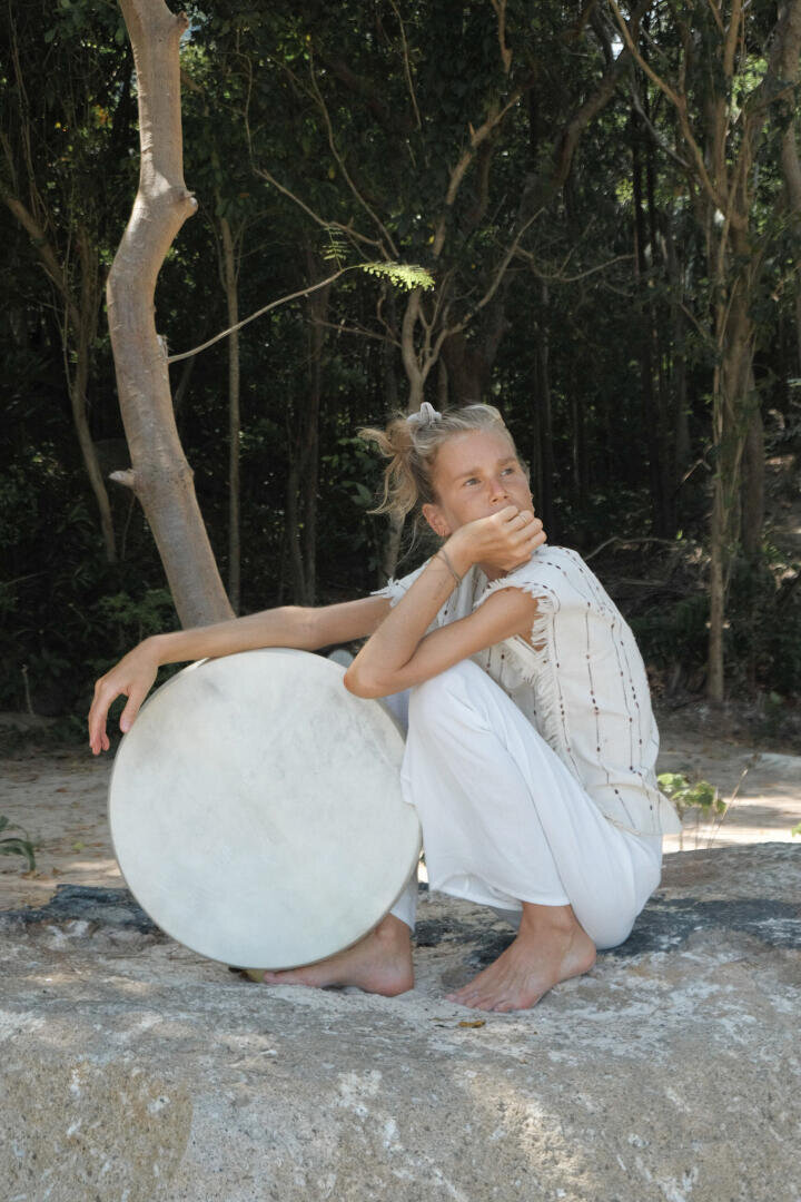 Eine barfüßige, weiß gekleidete Person sitzt im Freien auf einem Felsen und hält eine große runde Trommel in der Hand.