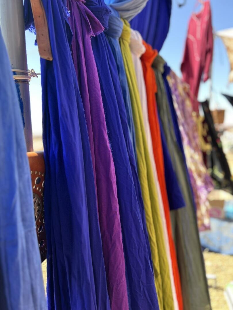 Bunte Stoffschals hängen vertikal in einer Reihe, darunter in Blau-, Violett-, Gelb-, Weiß- und Orangetönen, vor einem verschwommenen Hintergrund aus anderen Textilien und einem blauen Himmel.