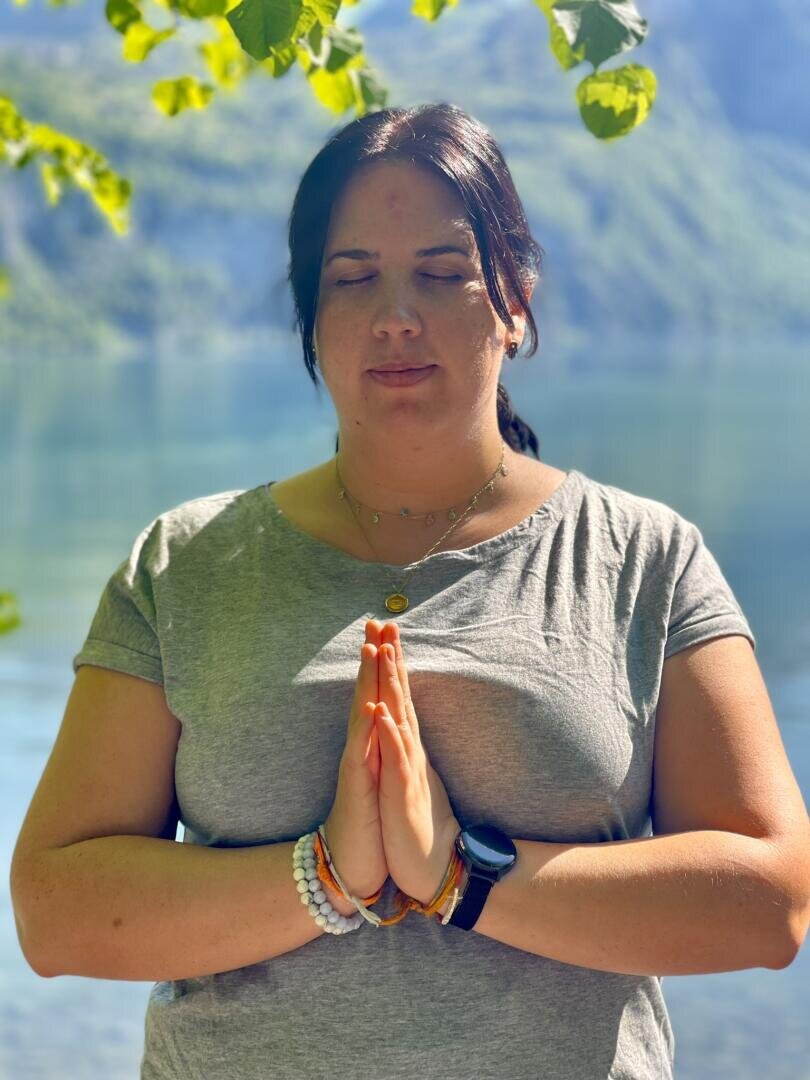 Eine Frau mit geschlossenen Augen steht im Freien in der Nähe eines Sees. Sie trägt ein graues Hemd, Armbänder und eine Uhr und hat die Hände in einer Gebets- oder Meditationshaltung zusammengepresst. Das Sonnenlicht fällt durch die grünen Blätter über ihr.