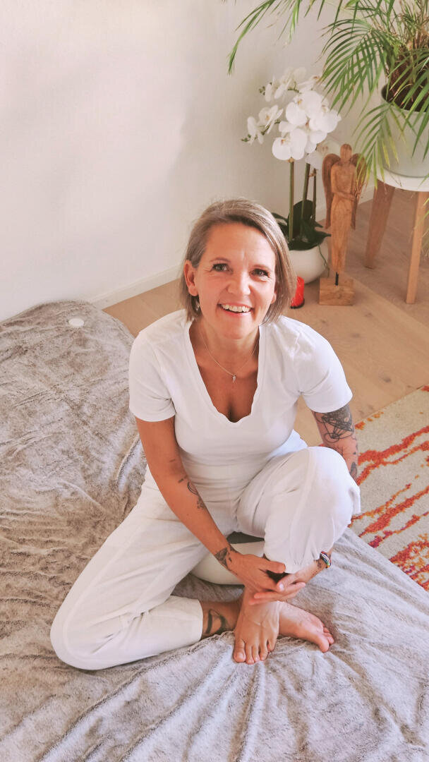 Eine lächelnde Frau mit kurzen Haaren und Tätowierungen sitzt im Schneidersitz auf einer Decke in einem hellen Raum mit Pflanzen, einer Engelsfigur und einem Blumenarrangement im Hintergrund. Sie trägt ein weißes Hemd und eine weiße Hose.