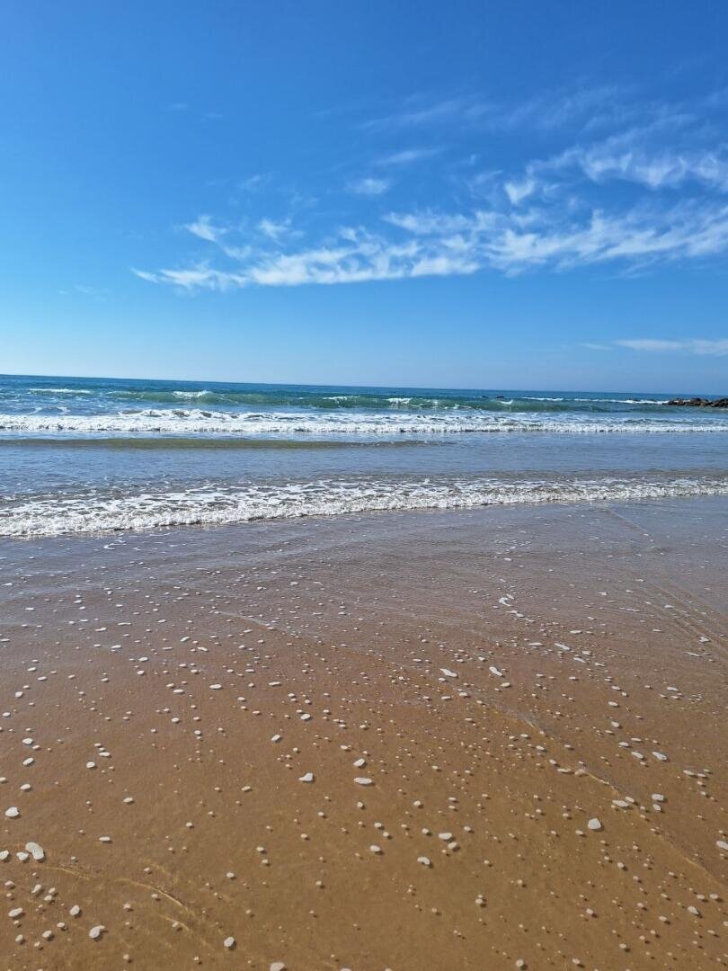Ein sonniger Strand, an dem sanfte Wellen über den nassen Sand rollen; leichter Schaum verteilt sich am Ufer. Der Himmel ist strahlend blau, und über dem ruhigen Meereshorizont ziehen zarte weiße Wolken dahin.