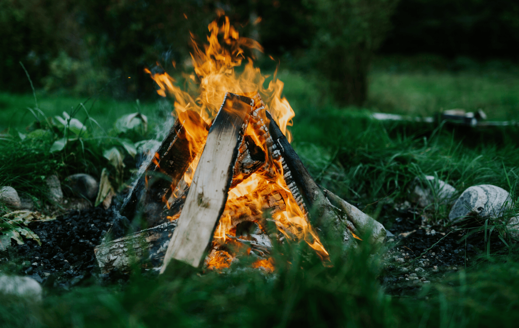 Ein Lagerfeuer brennt hell mit orangefarbenen Flammen und Rauch, umgeben von grünem Gras und Steinen in einer Außenumgebung. Holzscheite sind in Form eines Tipis angeordnet, und im unscharfen Hintergrund sind Bäume zu sehen.