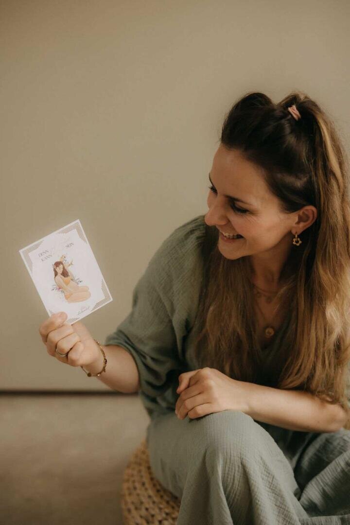 Eine lächelnde Frau mit langen braunen Haaren und grünem Outfit sitzt da und hält eine Karte mit einer illustrierten Frau und Text hoch. Der Hintergrund ist neutral und unaufgeregt.