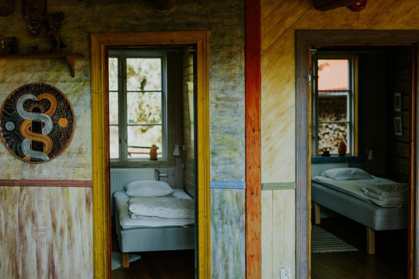Von einem farbenfrohen, rustikalen Flur aus gelangt man durch Türöffnungen in zwei gemütliche Schlafzimmer mit Einzelbetten. Durch die Fenster in beiden Zimmern fällt Sonnenlicht, und an der Flurwand hängt ein dekoratives rundes Wandbild mit einem Schlangenmotiv.