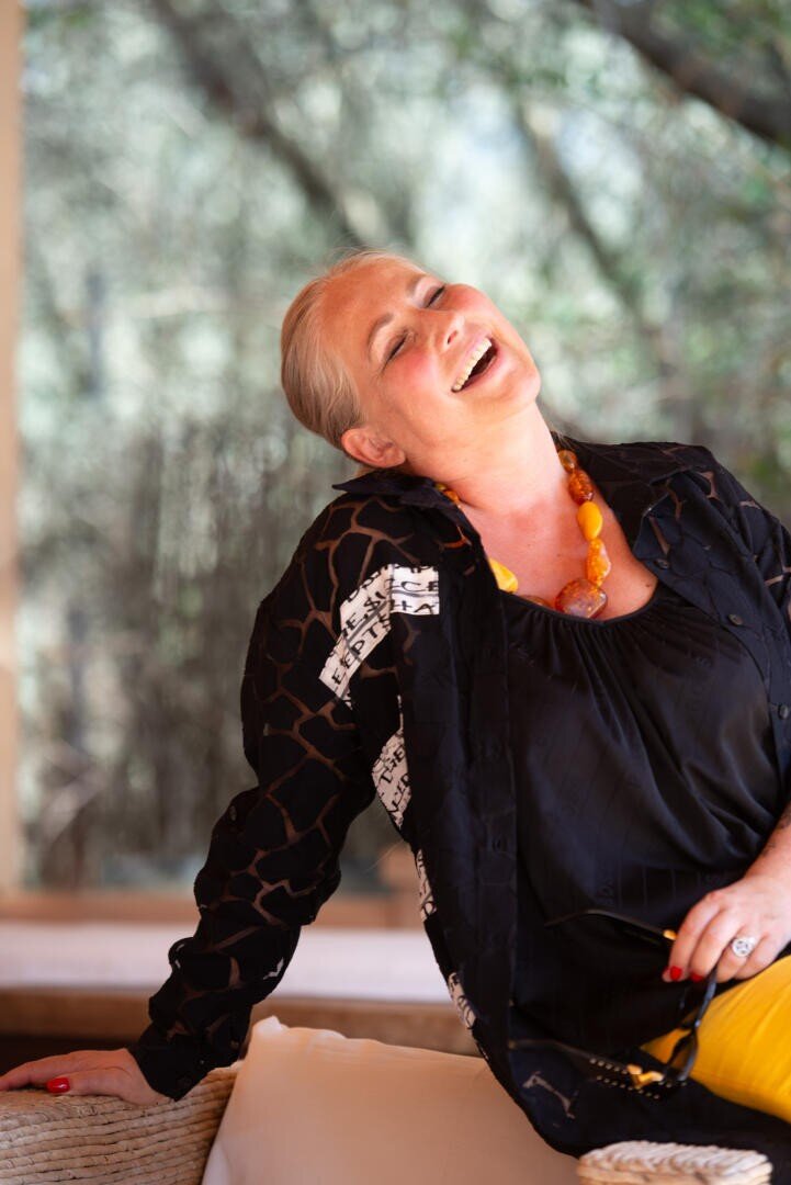 Eine Frau mit grauen Haaren, die ein schwarzes Hemd, eine gemusterte Jacke und eine klobige orangefarbene Halskette trägt, sitzt auf einer Couch im Freien und lehnt sich lächelnd und voller Freude zurück.