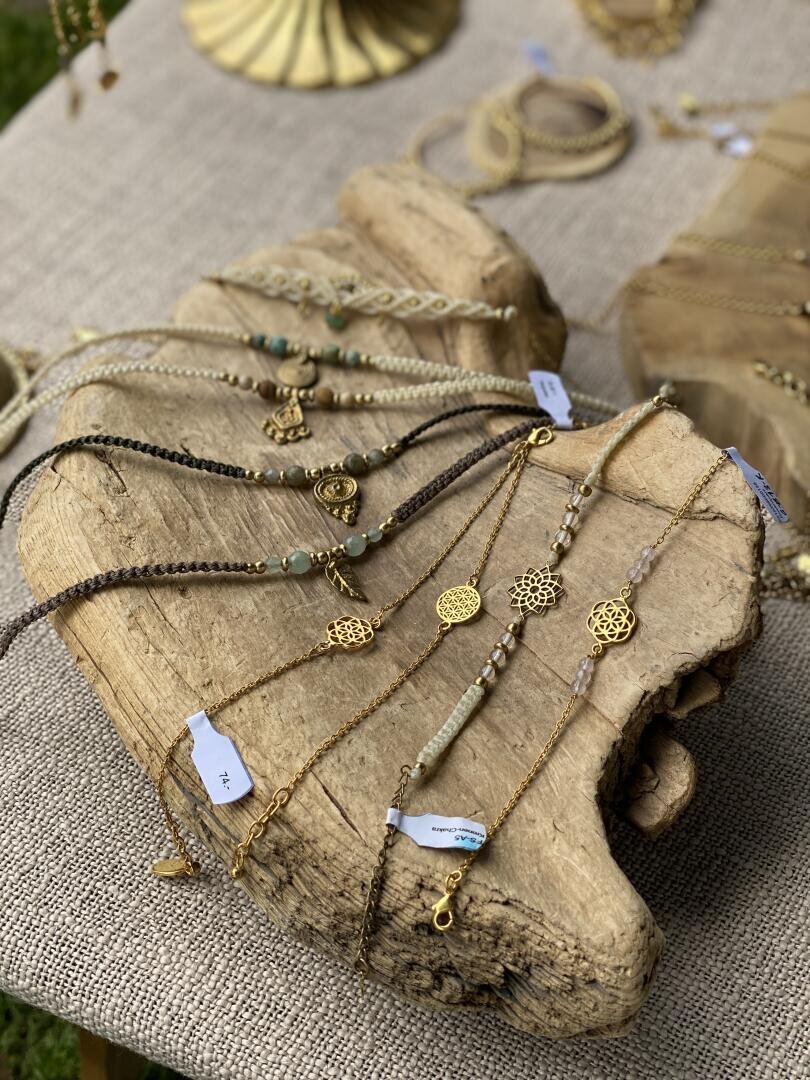 Handgefertigte Armbänder mit goldenen Anhängern und Perlen, präsentiert auf einem strukturierten Holzständer vor einem Jute-Stoff. Preisschilder sind zu sehen. Im Hintergrund sind weitere Schmuckstücke zu sehen.