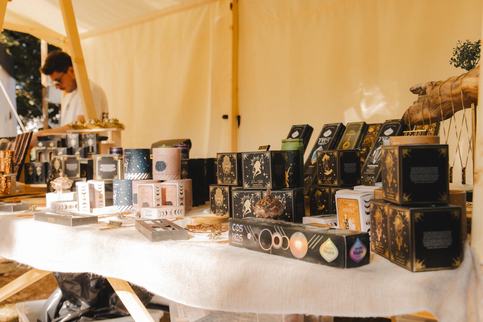Auf einem Marktstand sind ordentlich aufgereihte Schachteln mit Tee und anderen verpackten Waren auf einem Tisch ausgestellt. Hinter dem Tisch steht eine Person, die teilweise unscharf abgebildet ist. Der Stand ist unter einem weißen Vordach aufgebaut.