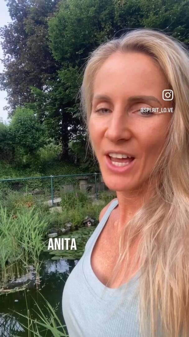 Eine Frau mit langen blonden Haaren und einem hellblauen Tank-Top steht im Freien in der Nähe eines Teiches mit Grün und Bäumen im Hintergrund. Der Text ANITA und ein Instagram-Symbol erscheinen auf dem Bild.