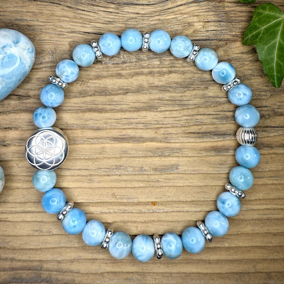 Ein blaues Perlenarmband mit silbernen Zwischenstücken und einem dekorativen silbernen Anhänger ist kreisförmig auf einer Holzunterlage angeordnet, daneben liegen ein blauer Stein und ein grünes Blatt.