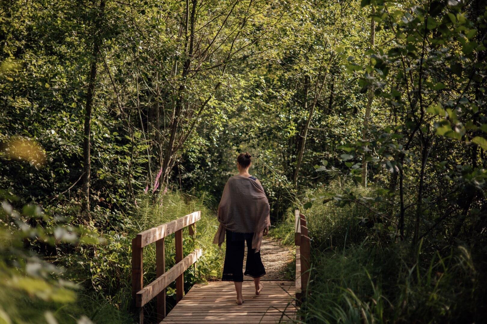 An einem sonnigen Tag läuft eine Person, die in einen Schal gehüllt ist, barfuß über eine Holzbrücke, die von üppig grünen Bäumen und dichter Vegetation umgeben ist.