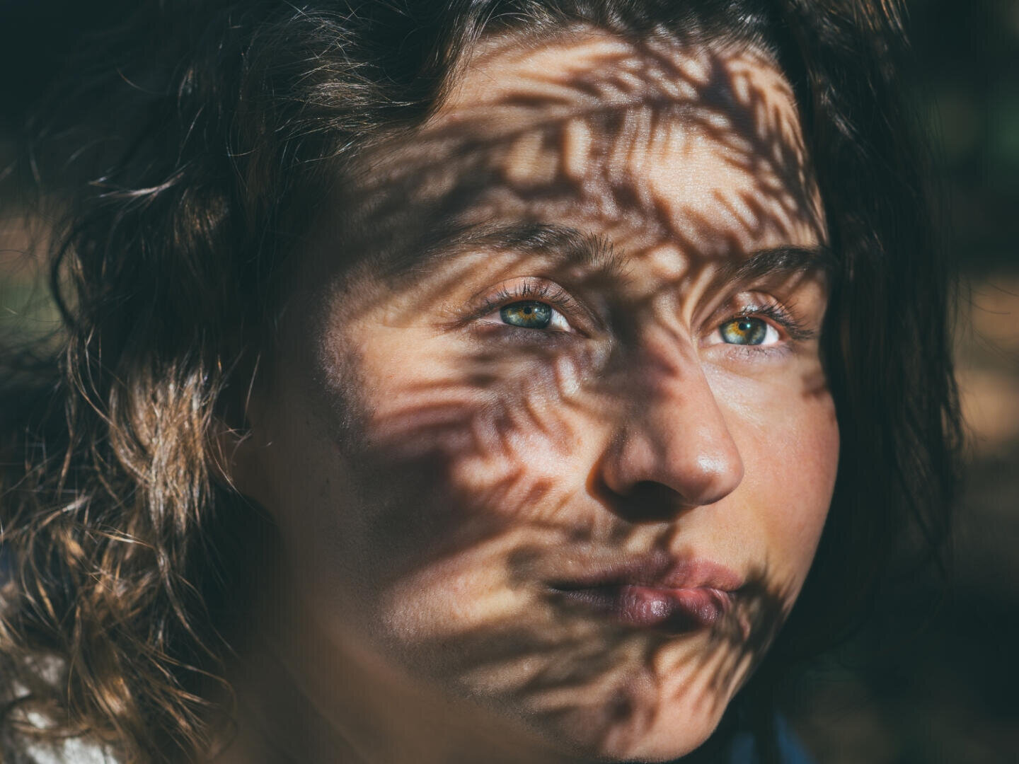 Eine Nahaufnahme des Gesichts einer Person, auf deren Haut das Sonnenlicht detaillierte, blattartige Schatten wirft. Die grünen Augen blicken leicht nach oben, umgeben von weichem, gewelltem Haar. Die Stimmung ist kontemplativ und künstlerisch.