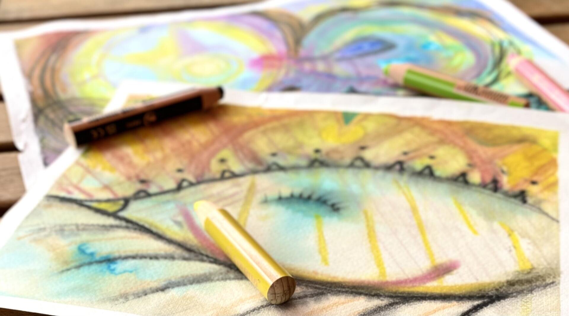 Zwei farbenfrohe abstrakte Zeichnungen auf Papier, die mit Pastellstiften entstanden sind. Auf dem Kunstwerk, das durch leuchtende Wirbel und Formen besticht, liegen mehrere Pastellstifte verstreut. Das Bild wirkt weich und leicht unscharf.