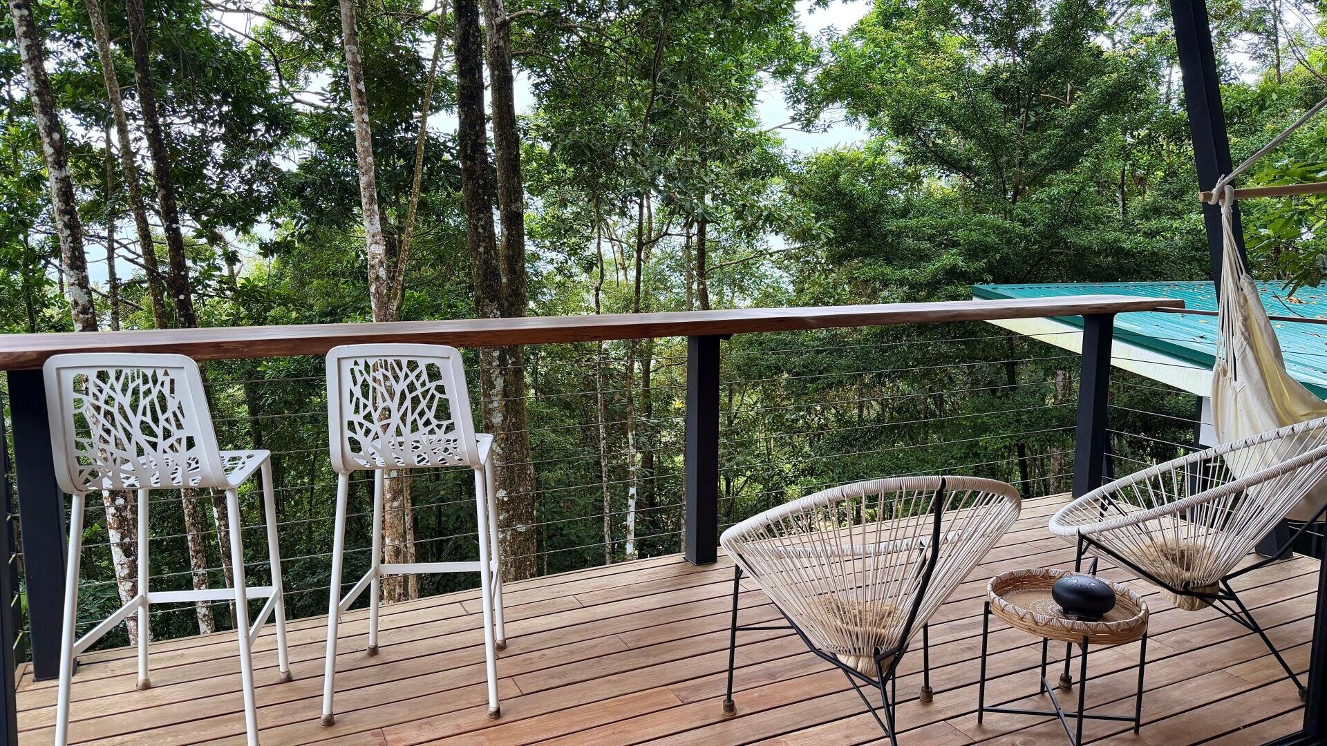 Eine Holzterrasse mit Blick auf einen üppigen Wald, mit zwei modernen weißen Barhockern an einem Geländer und zwei Drahtstühlen um einen kleinen runden Tisch. An der Seite hängt eine Hängematte, die für eine entspannte Atmosphäre im Freien sorgt.
