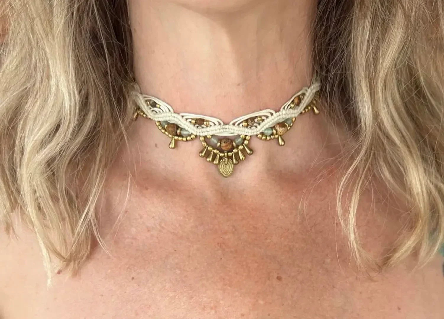 Eine Frau mit welligem blondem Haar trägt ein handgefertigtes Halsband mit cremefarbenen Fäden und goldenen Perlen, das an ihrem Hals und auf ihrer oberen Brust aufliegt.