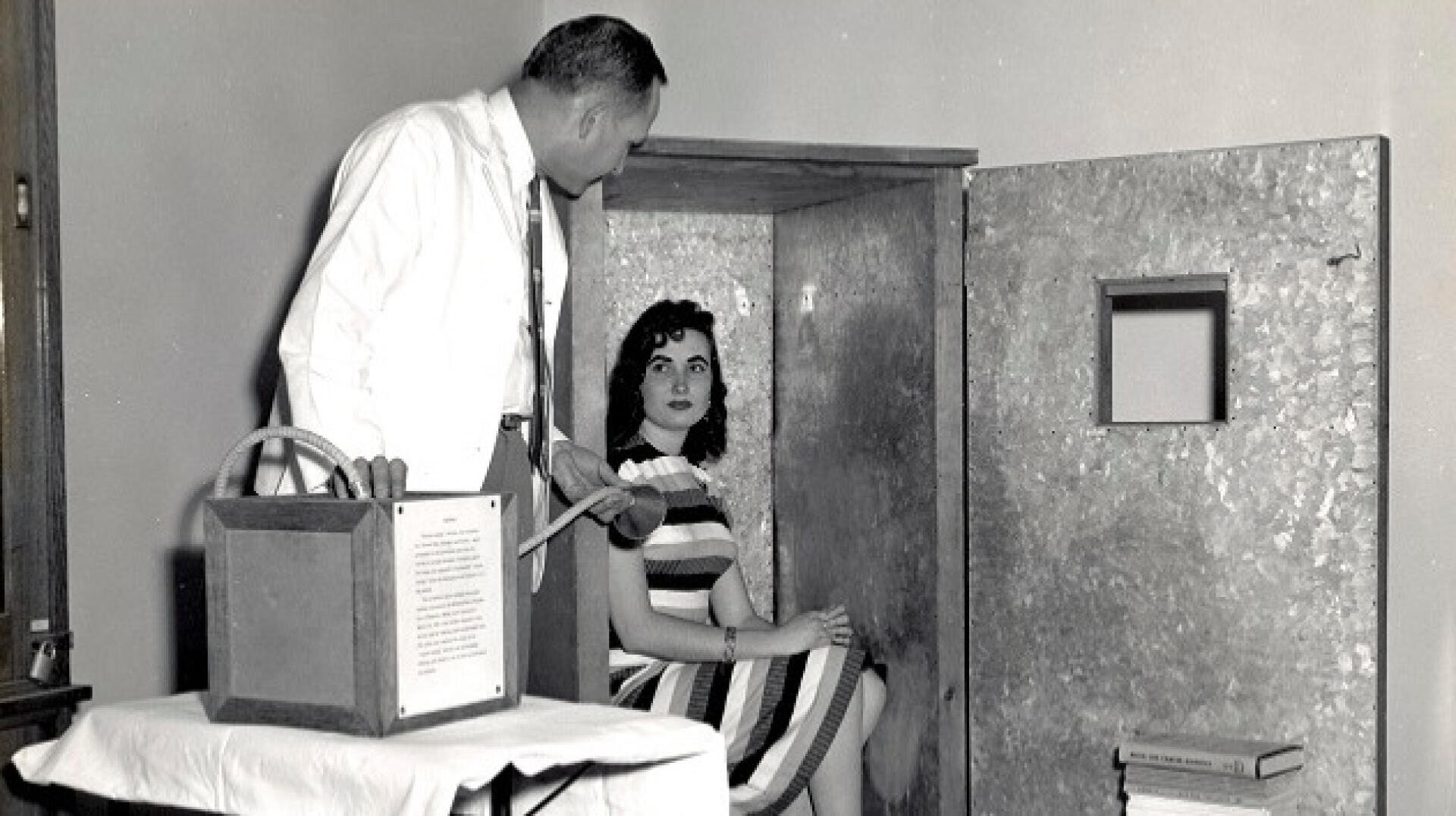 Eine Frau sitzt in einem kleinen Metallkasten mit einem quadratischen Fenster, während ein Mann in Hemd und Krawatte in der Nähe steht und ein Gerät mit einem Schlauch bedient, der an dem Kasten befestigt ist.