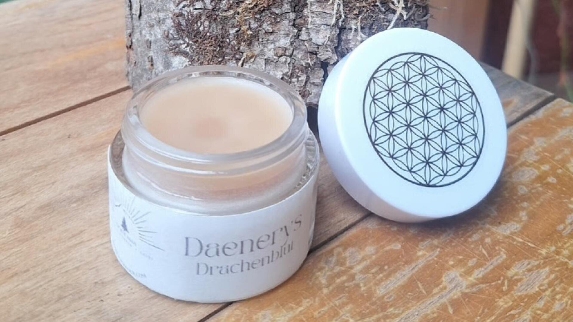 Ein Glasgefäß mit der Aufschrift „Daeners Drachenblut“ steht offen auf einer Holzfläche. Der weiße Deckel mit einem geometrischen, blumenähnlichen Muster liegt daneben. Im Hintergrund ist ein Baumstamm zu sehen.
