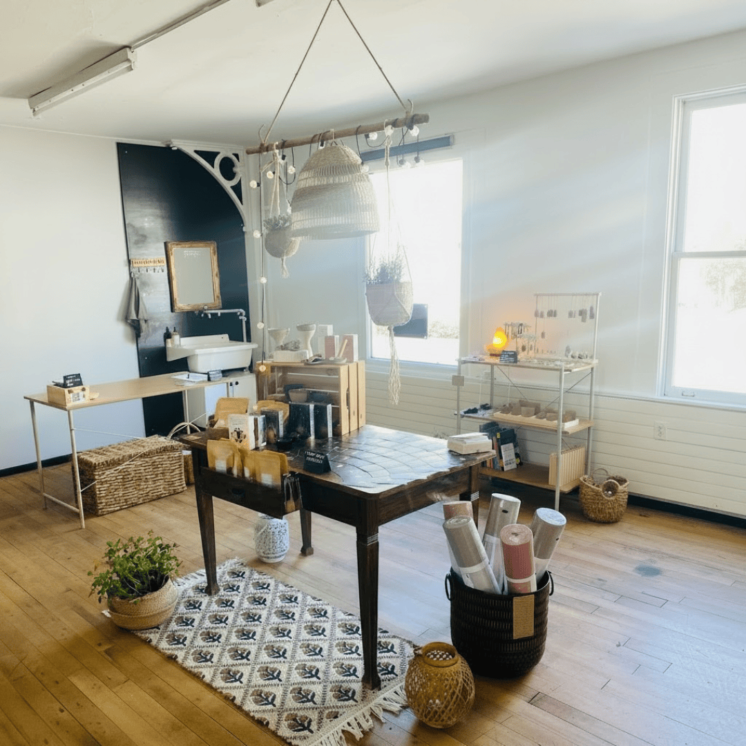 Eine sonnendurchflutete Boutique mit Holzböden, einem Tisch, auf dem Wellnessprodukte, geflochtene Körbe, Yogamatten und Pflanzen ausgestellt sind. Ein Waschbecken, Teppiche und Regale mit verschiedenen Artikeln sind zu sehen und schaffen eine gemütliche, einladende Atmosphäre.
