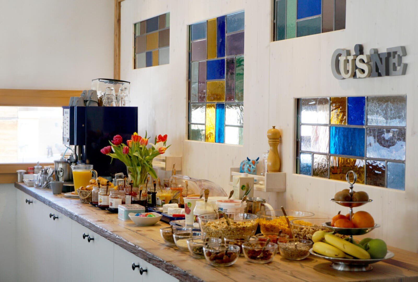 Eine lange Theke mit verschiedenen Frühstücksprodukten, Gläsern, Obst, Saft, Gebäck und Müsli. Es gibt eine Kaffeemaschine, bunte Glasfenster, Blumen in einer Vase und Küchendekoration an der Wand.