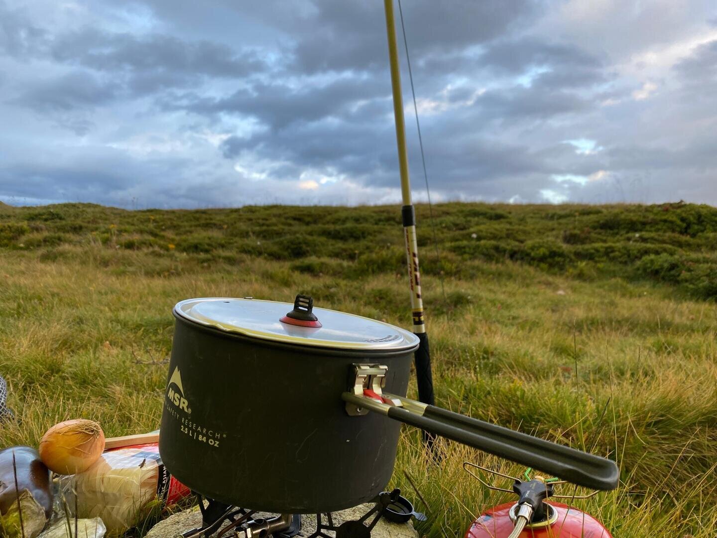 Ein Campingkocher mit einem Topf darauf steht im hohen Gras. Daneben steht eine Angelrute, und im Hintergrund erstreckt sich ein bewölkter Himmel über eine grasbewachsene Landschaft.