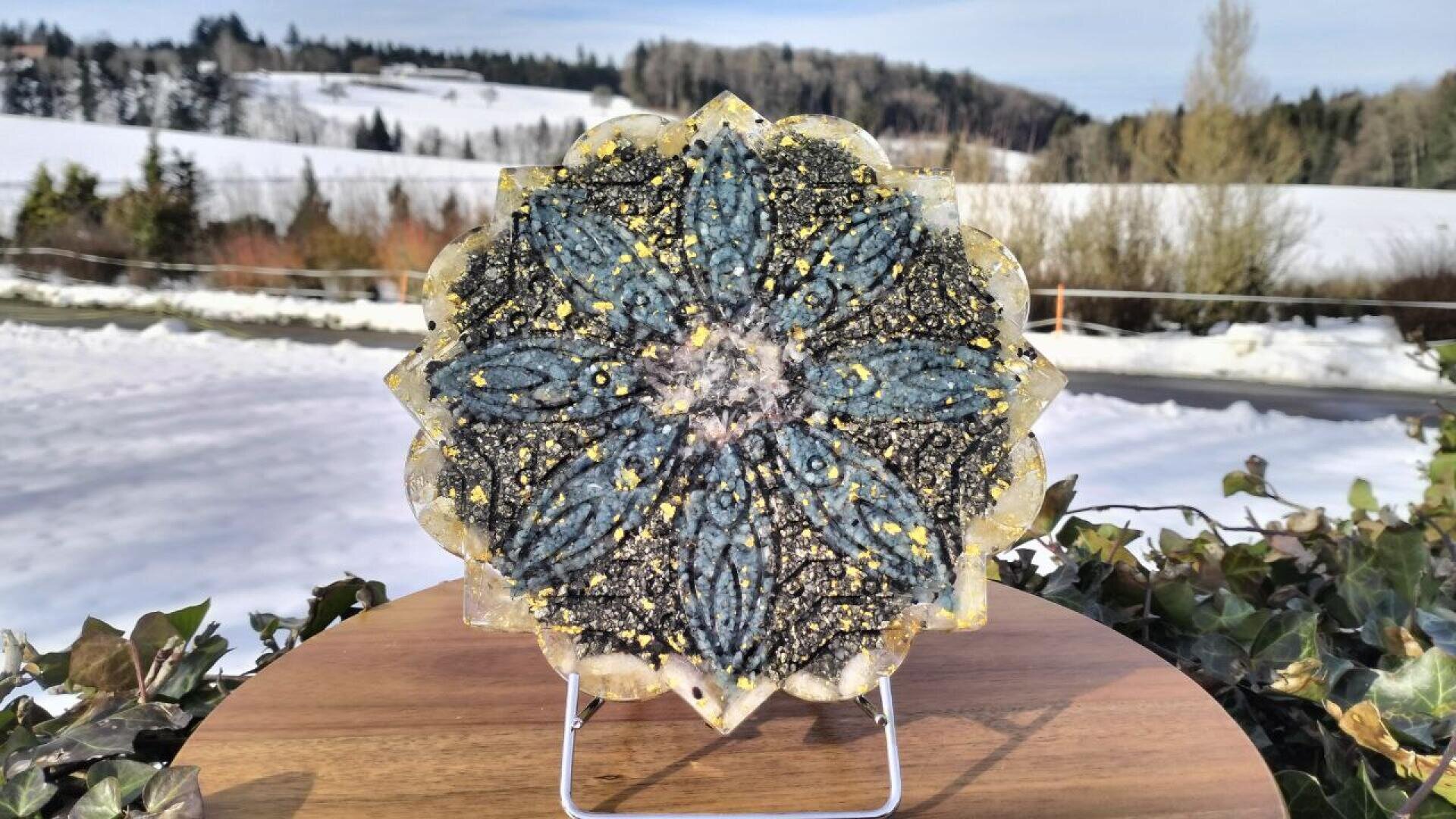 Ein dekoratives, blumenförmiges Kunstwerk aus Harz mit blauen und goldenen Akzenten wird auf einem Holzständer im Freien ausgestellt, mit einer verschneiten Landschaft und Bäumen im Hintergrund.