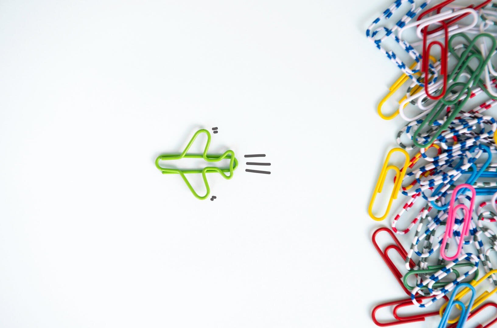 Eine grüne Büroklammer in Form eines Flugzeugs befindet sich auf einem weißen Hintergrund mit drei schwarzen Linien, die die Bewegung darstellen sollen. Auf der rechten Seite sind bunte Standard- und gestreifte Büroklammern auf einem Haufen verstreut.